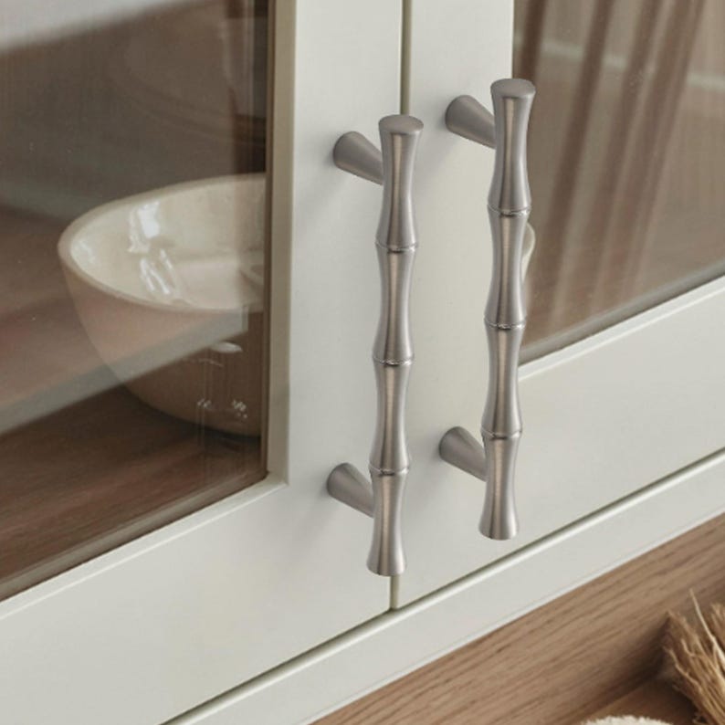 May include: Two vertical cabinet handles with a brushed silver finish and a segmented bamboo-like texture. Each handle is mounted to a white cabinet door frame with glass panels, featuring a smooth, metallic surface and rounded ends.