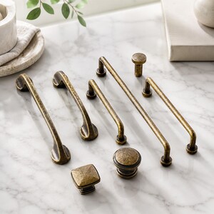 May include: A collection of antique brass cabinet hardware including two long curved handles, two straight bar pulls, one small cylindrical knob, one round knob, and one square knob, all featuring a weathered, textured metallic finish.
