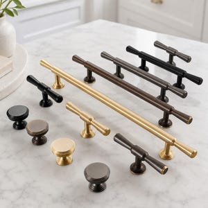 Op de afbeelding: Een collectie meubelbeslag bestaande uit diverse T-vormige handgrepen en ronde knoppen in geborsteld goud, matzwart en donkerbrons. De onderdelen zijn voorzien van gladde cilindrische stangen, taps toelopende uiteinden en ronde montagevoeten.