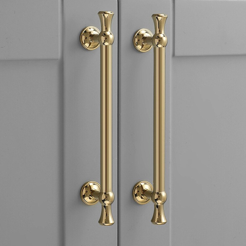 K&ouml;nnte beinhalten: Zwei vertikale Schrankgriffe mit poliertem Gold-Finish. Jeder Griff besteht aus einer langen, glatten zylindrischen Stange mit dekorativen abgerundeten Endst&uuml;cken oben und unten, befestigt durch zwei kreisf&ouml;rmige Montageplatten.