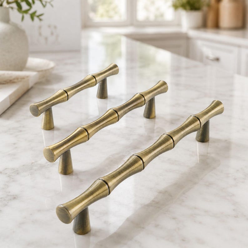 May include: Three cabinet pulls with a metallic antique brass finish. Each handle features a segmented design resembling bamboo stalks, with two conical mounting posts supporting the horizontal bar. The hardware has a smooth, polished surface.