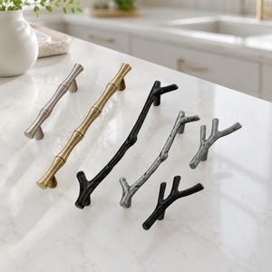 May include: A collection of metal cabinet hardware including two straight bamboo-style pulls in silver and gold, and four branch-shaped pulls in matte black and textured silver finishes, arranged diagonally on a white marble surface.