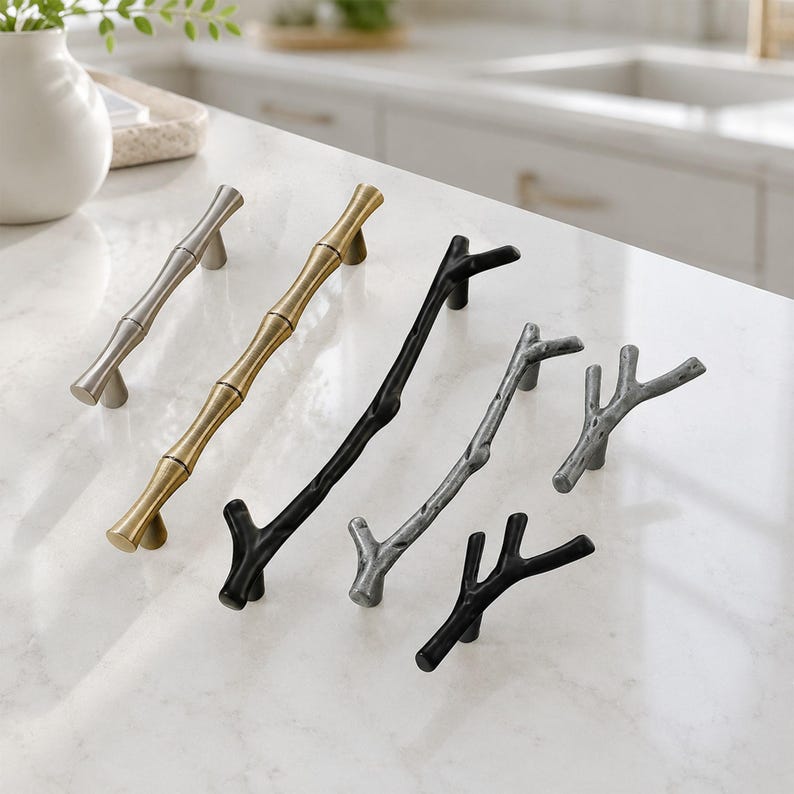 May include: A collection of metal cabinet hardware including two straight bamboo-style pulls in silver and gold, and four branch-shaped pulls in matte black and textured silver finishes, arranged diagonally on a white marble surface.