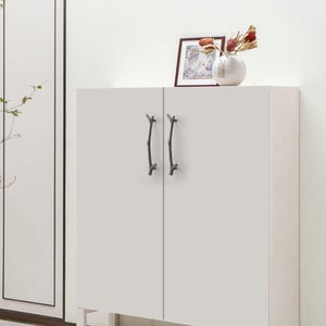 May include: A white cabinet featuring two dark gray metal door handles shaped like tree branches. The cabinet has a flat, smooth surface with vertical doors. A small framed picture and a white vase with dried flowers sit on top of the cabinet.