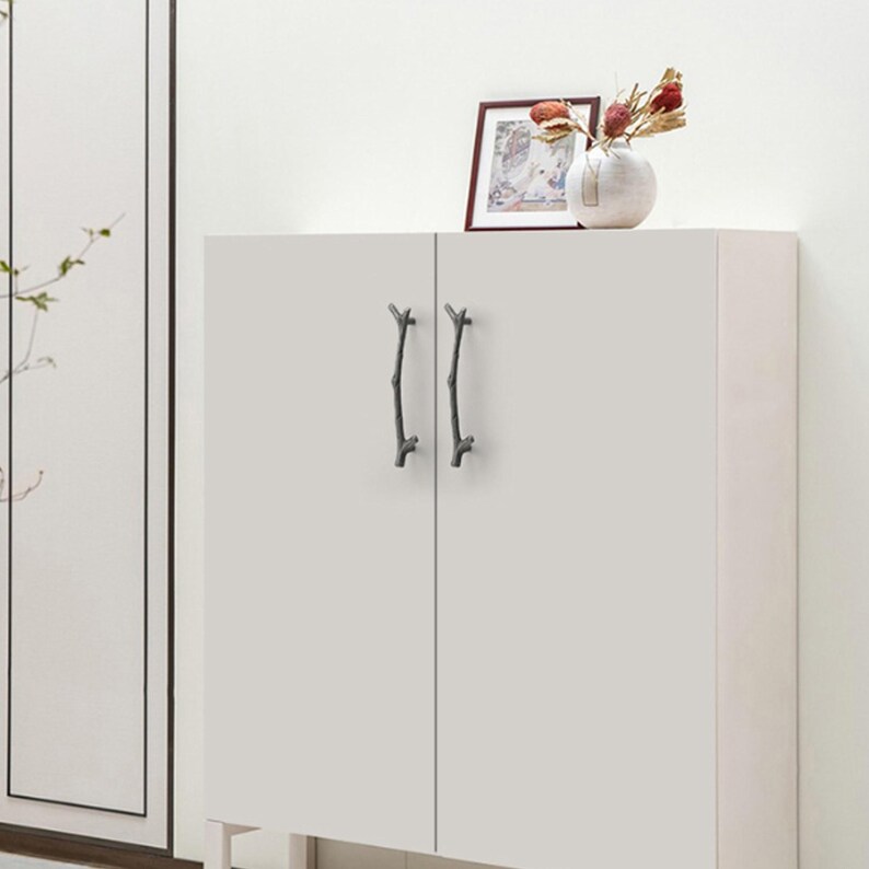 May include: A white cabinet featuring two dark gray metal door handles shaped like tree branches. The cabinet has a flat, smooth surface with vertical doors. A small framed picture and a white vase with dried flowers sit on top of the cabinet.