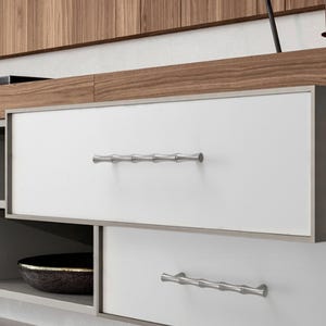 May include: A set of two horizontal drawer pulls with a metallic silver finish and a segmented bamboo-like texture. These hardware pieces are mounted onto white rectangular drawer fronts that are framed by a thin, dark-toned metal border.