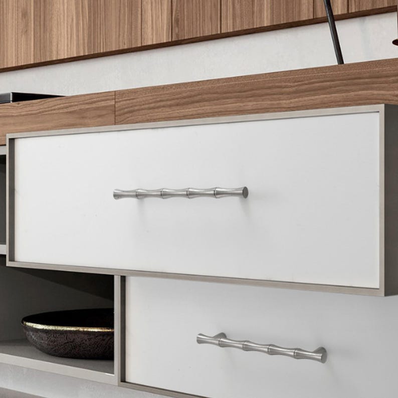 May include: A set of two horizontal drawer pulls with a metallic silver finish and a segmented bamboo-like texture. These hardware pieces are mounted onto white rectangular drawer fronts that are framed by a thin, dark-toned metal border.