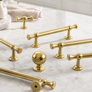 May include: A collection of polished gold-tone metal cabinet hardware, including several long bar pulls with cylindrical ends and a single spherical knob. Each piece features a smooth, reflective finish and a sturdy base for mounting to furniture.