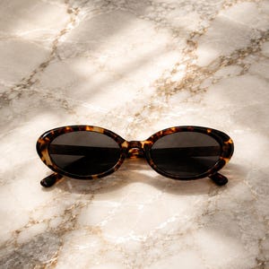 Peut inclure: Lunettes de soleil ovales dotées d'une monture à motif écaille de tortue marron et noir avec des verres teintés foncés. Ces lunettes présentent une forme arrondie classique avec un pont fin reliant les deux verres et des branches incurvées.