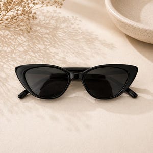 Könnte beinhalten: Schwarze Cat-Eye-Sonnenbrille mit glänzendem Kunststoffrahmen und dunkel getönten Gläsern. Die Brille hat ein Design mit spitz zulaufenden Außenkanten und einen schmalen Steg, der die beiden ovalen Gläser verbindet, was für ein einheitliches Aussehen sorgt.
