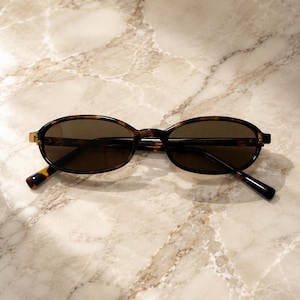 May include: Oval sunglasses featuring a brown and black tortoiseshell pattern frame with dark tinted lenses. The glasses have thin, curved temples and a bridge connecting the two oval-shaped rims, providing a classic look with a polished finish.