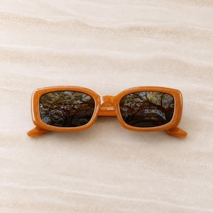 Peut inclure: Lunettes de soleil rectangulaires avec une monture ambrée brillante et des verres teintés foncés. Les montures présentent un design aux bords arrondis et lisses avec un pont reliant les deux verres. Les verres reflètent des arbres.