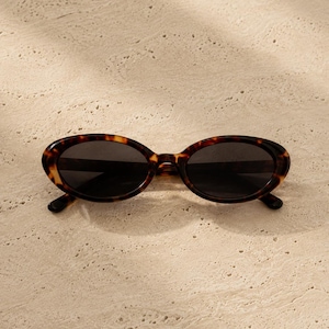 May include: Oval sunglasses featuring a dark brown and black tortoiseshell pattern frame with dark tinted lenses. The eyewear has a rounded, elongated shape with a bridge connecting the two lenses and curved temple arms extending from the sides.