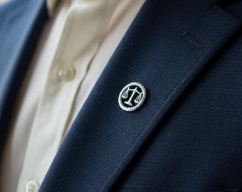 May include: A circular silver lapel pin featuring a black center with a raised silver scale of justice symbol, attached to the lapel of a dark navy blue blazer worn over a light cream-colored button-down shirt with a visible collar and buttons.