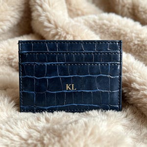 May include: A rectangular navy blue card holder featuring a textured crocodile skin pattern and the gold embossed letters KL centered on the front. The accessory has horizontal slots for cards and is constructed with visible stitching along the edges.