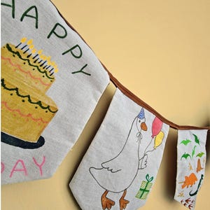 Op de afbeelding: Een stoffen vlaggenlijn met drie wimpels met handgetekende illustraties. De eerste toont een gele verjaardagstaart en de tekst HAPPY. De tweede toont een gans met een feesthoedje die ballonnen en een cadeau vasthoudt. De derde toont dinosaurussen.