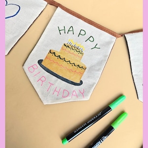 Op de afbeelding: Een crèmekleurige stoffen vlaggetje met een handgetekende illustratie van een gele verjaardagstaart met twee lagen en brandende kaarsjes. De tekst "HAPPY" staat in het groen boven de taart en "BIRTHDAY" in het roze eronder. Twee groene textielstiften.