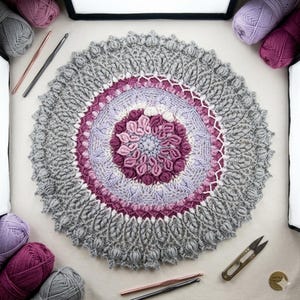 Könnte beinhalten: Ein rundes Häkelmandala mit komplexen Texturen in den Farben Grau, Lavendel und tiefem Magenta. Das Design umfasst schichtweise Blütenblattmuster, Noppenstiche und einen gewellten Rand, umgeben von Garnknäueln und verschiedenen Häkelnadeln.