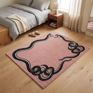 May include: A rectangular area rug in a dusty pink hue featuring a black graphic illustration of two snakes facing each other. The snakes are depicted with open mouths and coiled bodies, creating a border-like design across the centre of the rug.