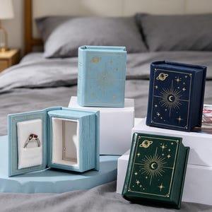 May include: Three book-shaped jewelry boxes in light blue, dark blue, and dark green, each featuring gold celestial foil stamping of a sun, moon, and stars. One light blue box is open, revealing a white velvet ring insert holding a silver ring.