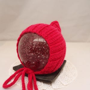 Könnte beinhalten: Eine rote Strickhaube mit Ohrenklappen bedeckt eine klare Glaskugel, die mit Blasen gefüllt ist. Die Haube hat einen Kordelverschluss und steht auf einem schwarzen quadratischen Sockel. Die Kugel steht auf einer weißen Spitzendeckchen.