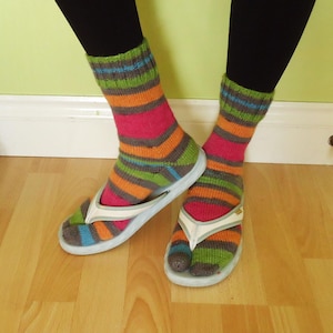 May include: Brightly coloured, striped socks in pink, orange, green, grey, and blue are worn with white and light blue flip-flops. The socks are pulled up over the ankles. The background is a light green wall and a wooden floor.