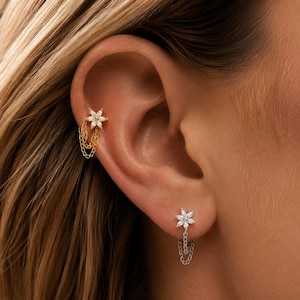 Könnte beinhalten: Ein goldfarbener Blumenohrring mit einer hängenden Kette am oberen Ohrknorpel und ein silberfarbener Blumenohrring mit einer hängenden Kette am Ohrläppchen, beide mit klaren, funkelnden Steinen in einer blumenförmigen Anordnung besetzt.