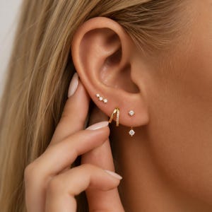 May include: A set of gold earrings consisting of a three-stone stud, a small hoop with a pavé band, and a single-stone stud with a dangling charm. The jewellery features clear, sparkling stones set in polished gold metal, creating a layered look.