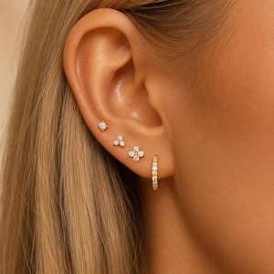 May include: A set of four gold-tone earrings worn in a single ear. The collection includes a small single-stone stud, a three-stone cluster stud, a four-stone flower-shaped stud, and a small gold hoop earring encrusted with clear white stones.