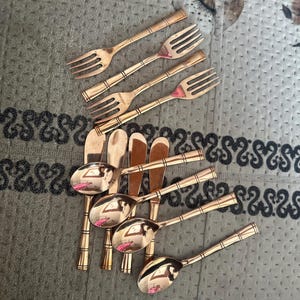 May include: A set of four forks and four spoons with a polished copper finish. Each piece of flatware features a slender handle with a series of horizontal bands or rings embossed along the stem, providing a consistent design across the collection.