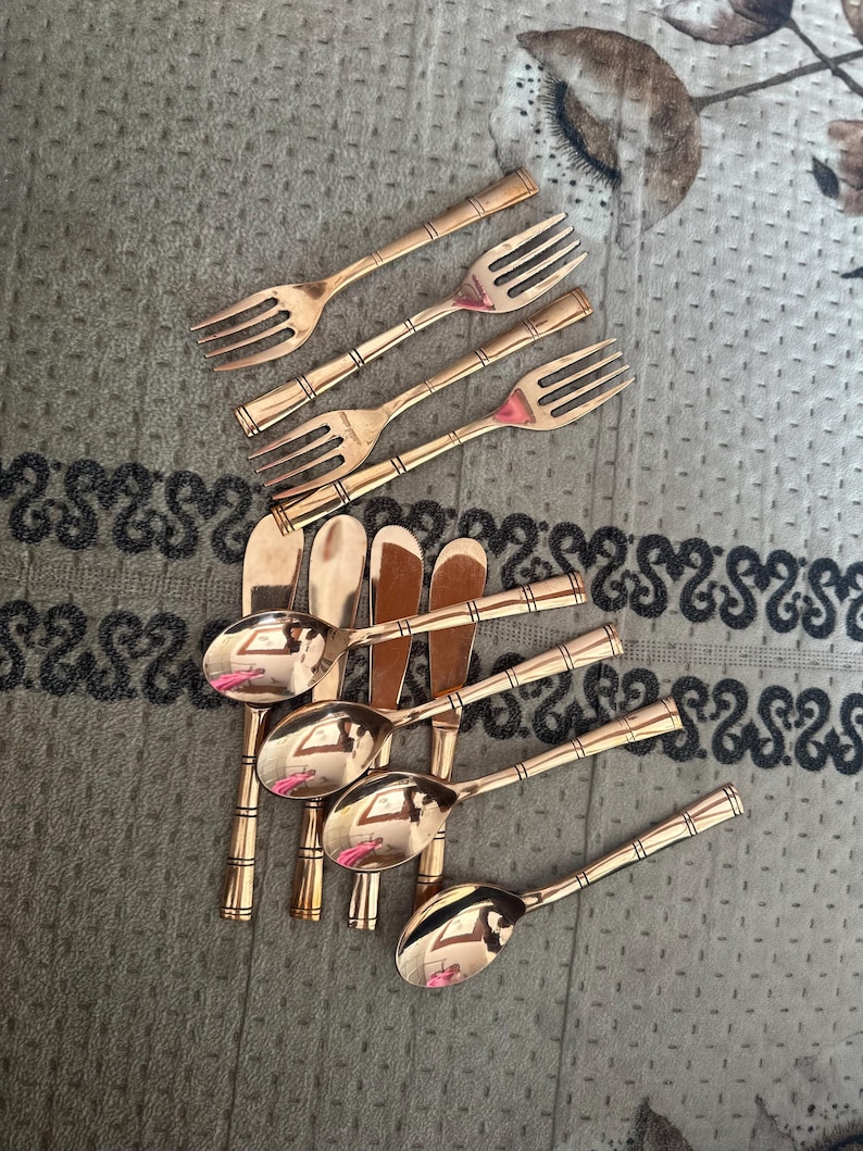 May include: A set of four forks and four spoons with a polished copper finish. Each piece of flatware features a slender handle with a series of horizontal bands or rings embossed along the stem, providing a consistent design across the collection.