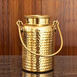 Könnte beinhalten: Ein zylindrischer, goldfarbener Metallbehälter mit einer gehämmerten Textur auf der Oberfläche. Der Behälter verfügt über einen glatten, flachen Rand an der Oberseite und einen geschwungenen, klappbaren Metallgriff, der an zwei seitlichen Ösen befestigt ist.