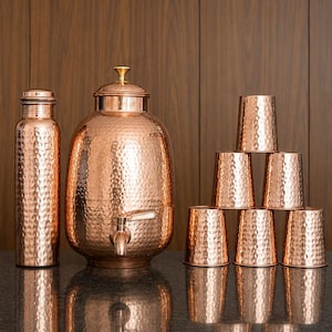 May include: A set of hammered copper drinkware including a tall cylindrical water bottle, a large round water dispenser with a metal spigot and lid, and six matching tumblers arranged in a pyramid stack, all featuring a textured finish.