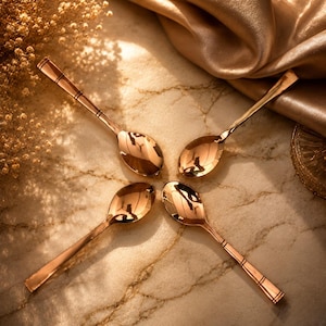 May include: Four polished copper-toned metal spoons arranged in a circular pattern. Each spoon features a smooth, reflective bowl and a slender handle with two distinct horizontal engraved lines near the end of the stem, creating a uniform set.