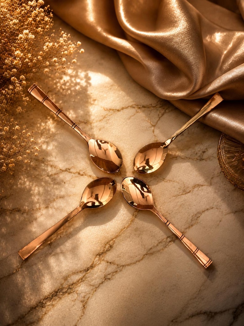 May include: Four polished copper-toned metal spoons arranged in a circular pattern. Each spoon features a smooth, reflective bowl and a slender handle with two distinct horizontal engraved lines near the end of the stem, creating a uniform set.