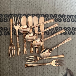 May include: A collection of rose gold-toned flatware featuring a bamboo-inspired design on the handles. The set includes several forks, spoons, and butter knives with a polished, reflective finish, arranged in a cluster on a textured surface.