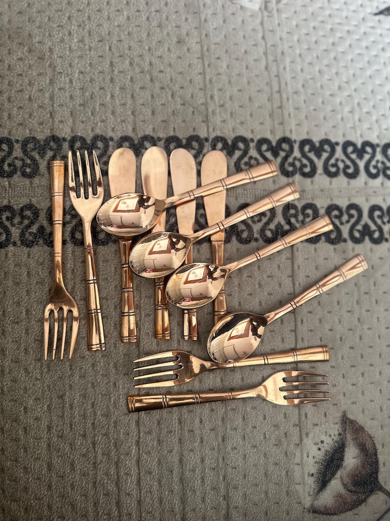 May include: A collection of rose gold-toned flatware featuring a bamboo-inspired design on the handles. The set includes several forks, spoons, and butter knives with a polished, reflective finish, arranged in a cluster on a textured surface.