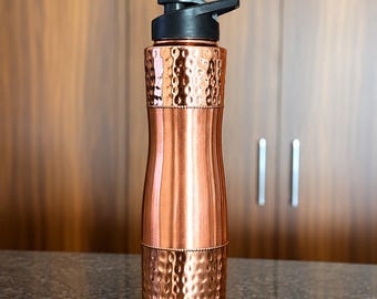 Peut inclure: Une bouteille d'eau de couleur cuivre avec une section centrale cylindrique lisse et des bandes texturées martelées en haut et en bas. La bouteille est dotée d'un couvercle en plastique noir avec un bec verseur et une poignée.