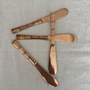 May include: Three copper-colored butter knives arranged in a cluster. Each knife features a smooth, rounded blade and a slender handle decorated with a series of thin, horizontal black lines etched into the metal surface at regular intervals.