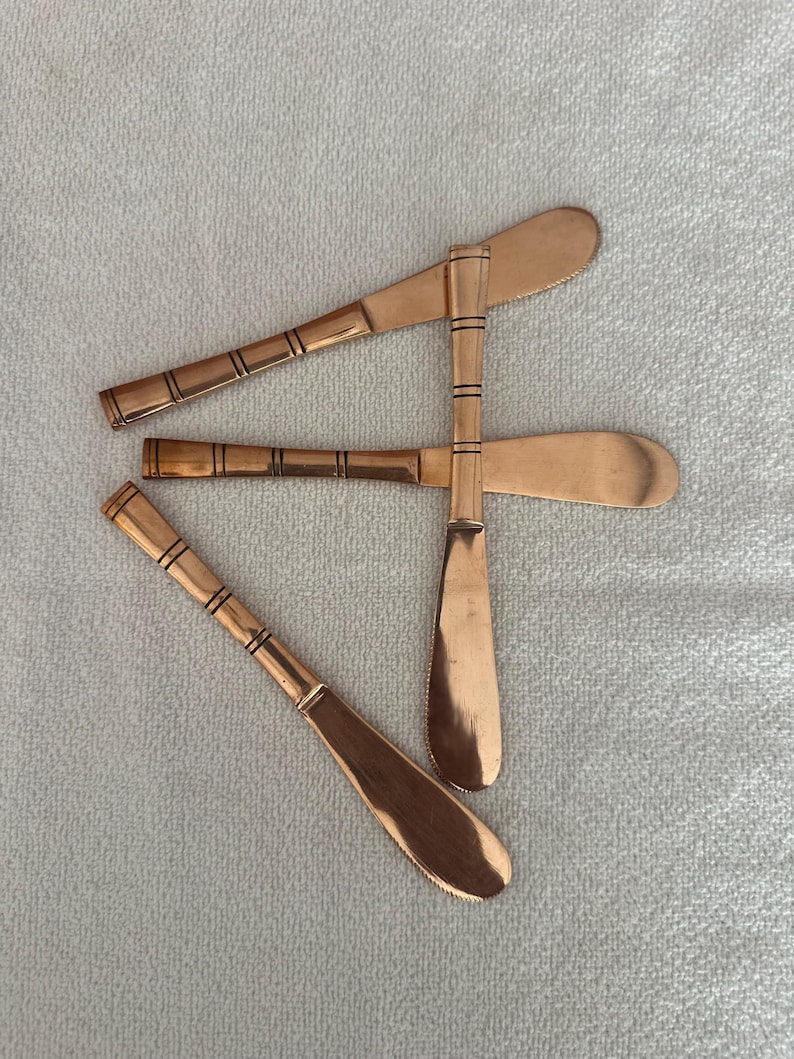 May include: Three copper-colored butter knives arranged in a cluster. Each knife features a smooth, rounded blade and a slender handle decorated with a series of thin, horizontal black lines etched into the metal surface at regular intervals.