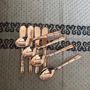 May include: A set of four copper-colored metal spoons with rounded bowls and handles featuring three horizontal engraved lines near the base. The spoons have a polished, reflective finish and are arranged in a staggered, overlapping formation.