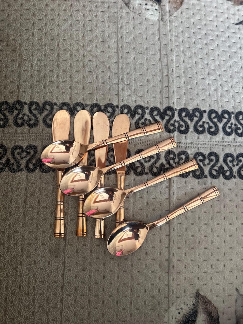 May include: A set of four copper-colored metal spoons with rounded bowls and handles featuring three horizontal engraved lines near the base. The spoons have a polished, reflective finish and are arranged in a staggered, overlapping formation.