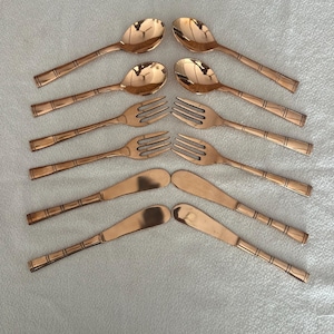 May include: A set of eight copper-colored flatware pieces arranged in two columns. The collection includes two spoons, two forks, and four butter knives, all featuring a smooth, reflective finish and a distinct banded design on the handles.