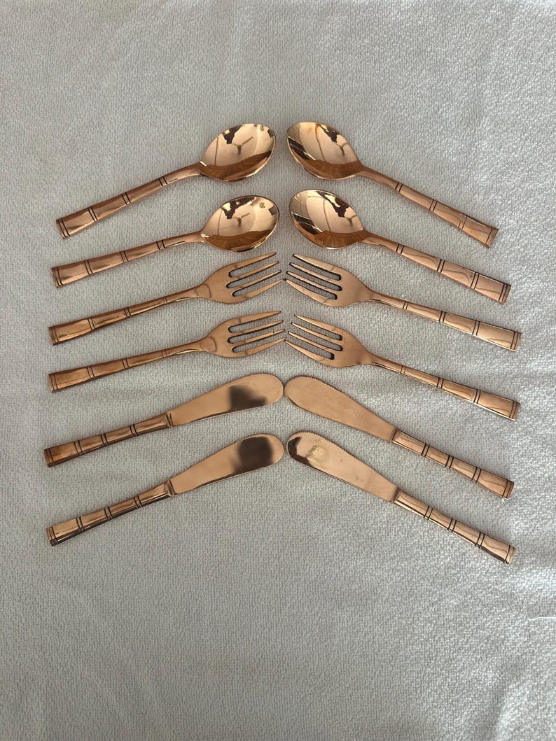 May include: A set of eight copper-colored flatware pieces arranged in two columns. The collection includes two spoons, two forks, and four butter knives, all featuring a smooth, reflective finish and a distinct banded design on the handles.