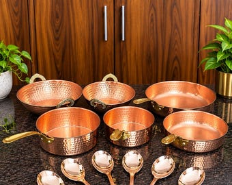 May include: A set of hammered copper cookware including two round bowls with side handles, three shallow pans with single handles, and six matching spoons. The cookware features a polished metallic finish and gold-toned hardware.