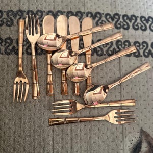 May include: A collection of copper-toned metal flatware including several forks and spoons with a bamboo-style handle design. The set features polished surfaces with horizontal line detailing on the handles, arranged in a loose cluster.