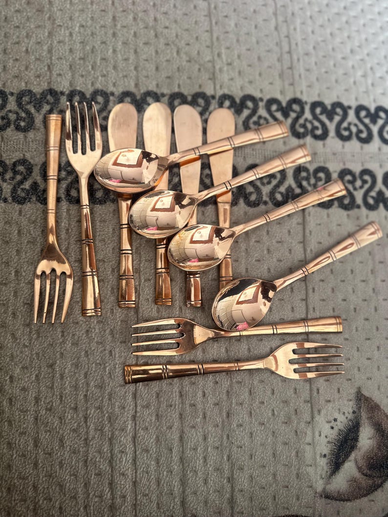 May include: A collection of copper-toned metal flatware including several forks and spoons with a bamboo-style handle design. The set features polished surfaces with horizontal line detailing on the handles, arranged in a loose cluster.