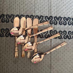 May include: A set of four copper-toned metal spoons with rounded bowls and slender handles. Each handle features a series of three horizontal engraved lines near the top and bottom, providing a consistent decorative detail across the collection.