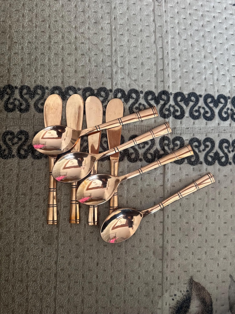 May include: A set of four copper-toned metal spoons with rounded bowls and slender handles. Each handle features a series of three horizontal engraved lines near the top and bottom, providing a consistent decorative detail across the collection.