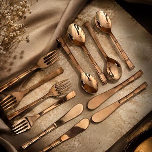 May include: A set of rose gold-toned flatware including three forks, two spoons, and three knives. Each piece features a polished metallic finish and a distinct handle design with three horizontal etched lines near the neck of the utensil.
